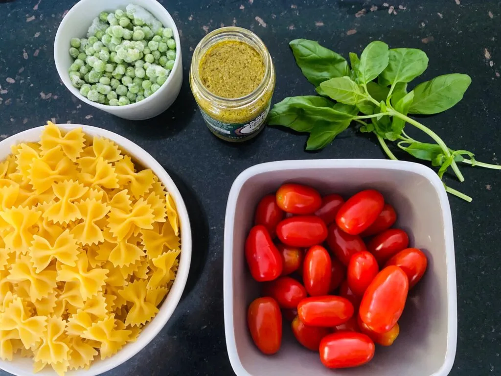 ingredients for pesto pasta salad, dry pasta, cherry tomatoes, fresh basil leaves, basil pesto, frozen peas