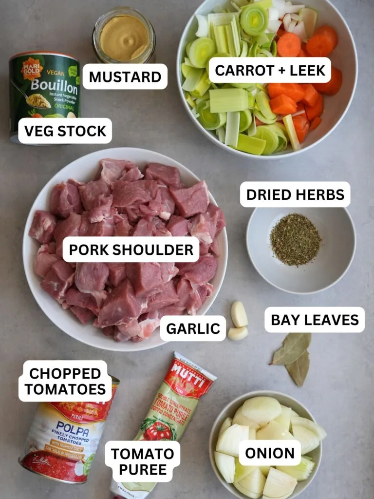 raw ingredients laid out and labelled for making a pork casserole in the slow cooker: vegetable stock, mustard, carrot and leek, pork shoulder, dried herbs, chopped tomatoes, garlic, bay leaves, tomato puree, onion