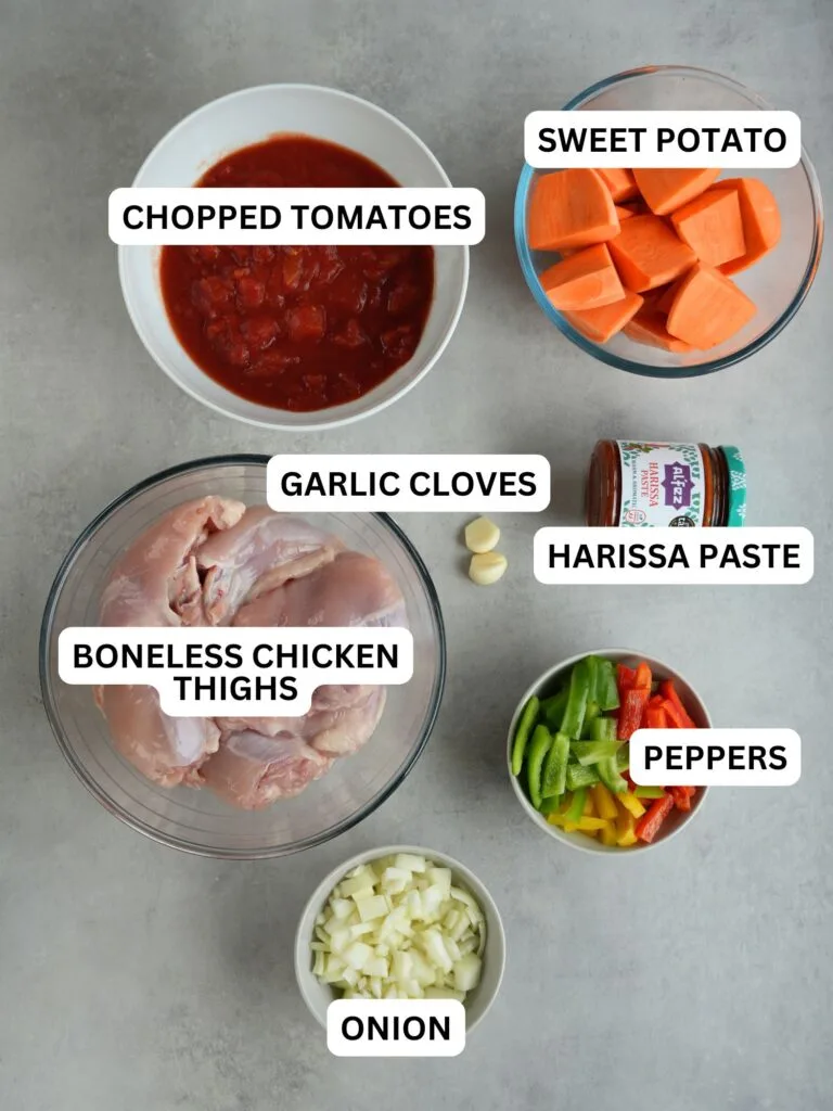 ingredients laid out for slow cooker chicken and sweet potato, labelled: tomatoes, sweet potatoes, chicken, garlic cloves, harissa paste, peppers, onion