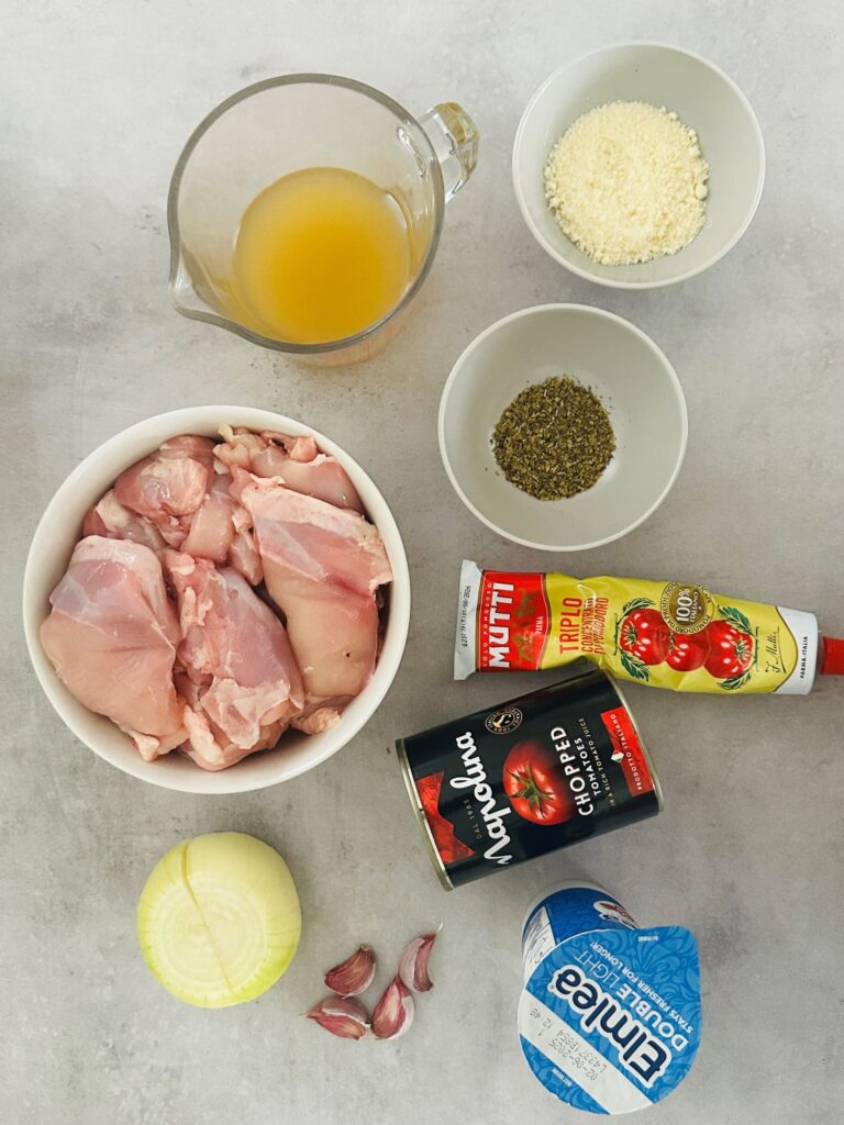 ingredients laid out for making tomato basil chicken in the slow cooker - raw chicken, chicken stock, dried basil, tomato puree, chopped tomatoes, onion, parmesan cheese, onion, 4 cloves garlic and cream
