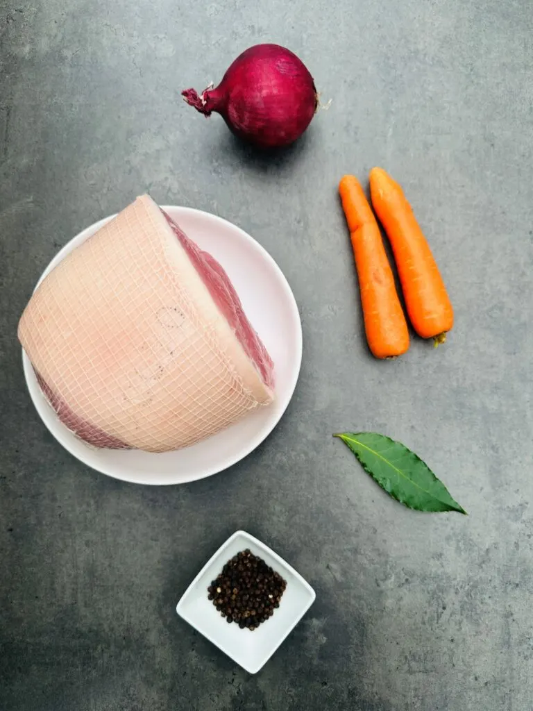 ingredients for cooking a gammon joint in a slow cooker: gammon joint, red onion, two carrots, bay leaf, peppercorns
