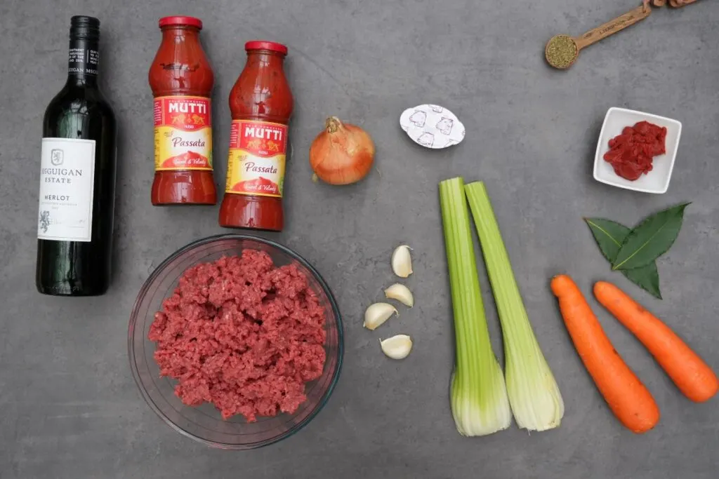 ingredients laid out for making ragu sauce for lasagne: beef mince, celery, carrots, garlic, tomato puree, beef stock, dried mixed herbs, onion, passata and red wine