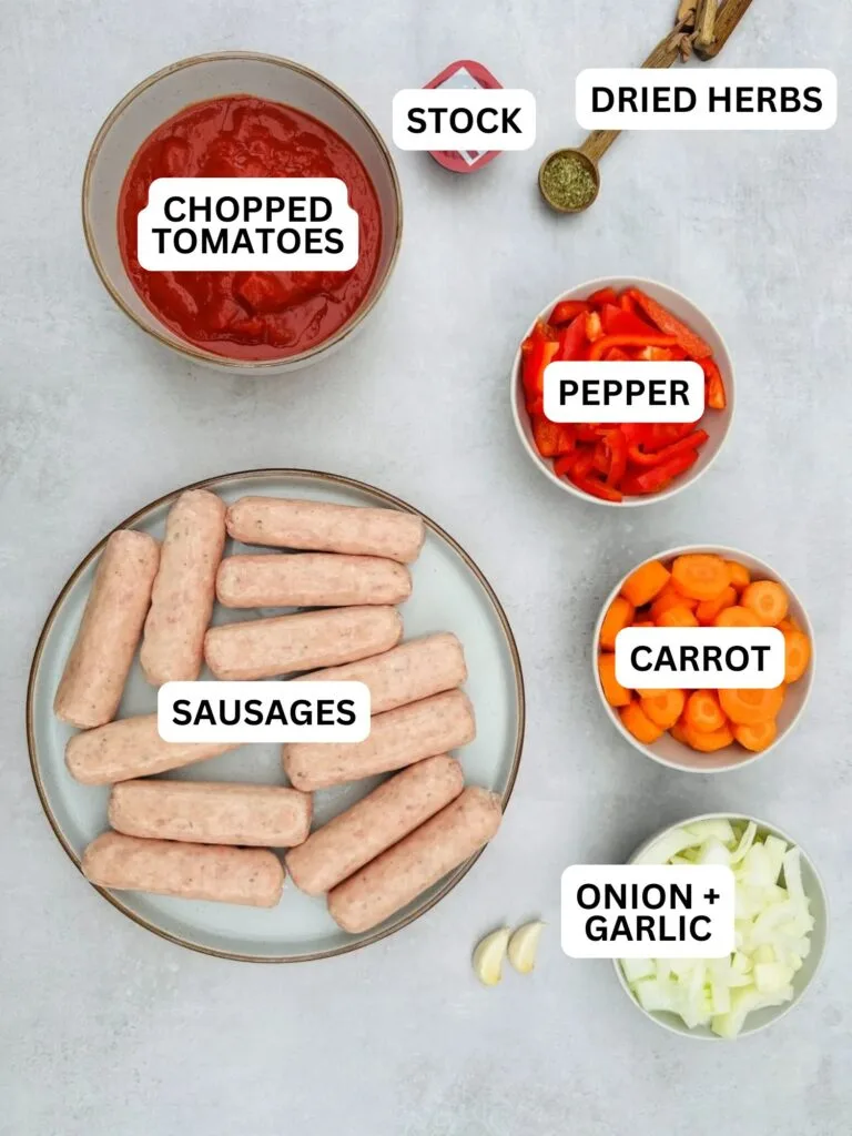 ingredients laid out for sausage casserole: 12 sausages on a plate, chopped carrots, chopped onion, chopped red pepper, 1 tsp dried mixed herbs, chopped tomatoes and beef stock pot