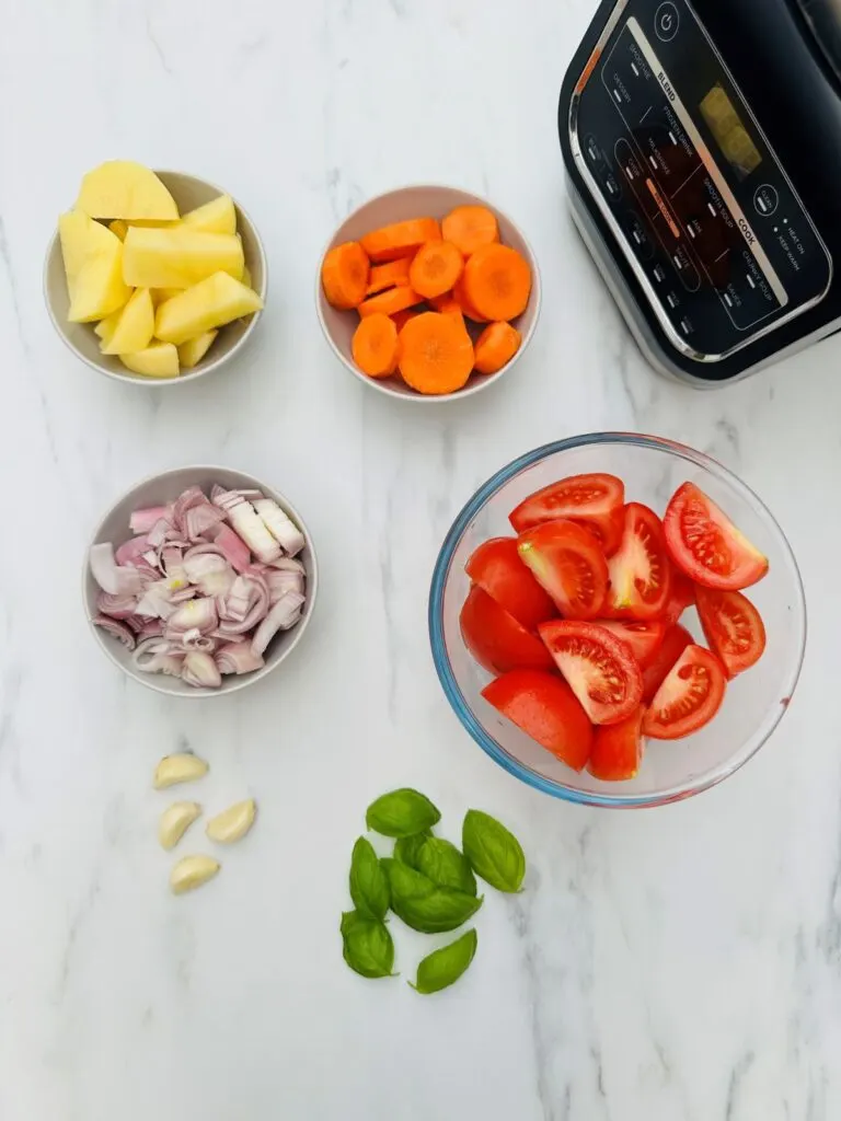 ingredients for tomato and basil soup in a soup maker: potatoes, tomatoes, carrots, shallots, garlic and basil leaves laid out next to a Ninja Soup Maker