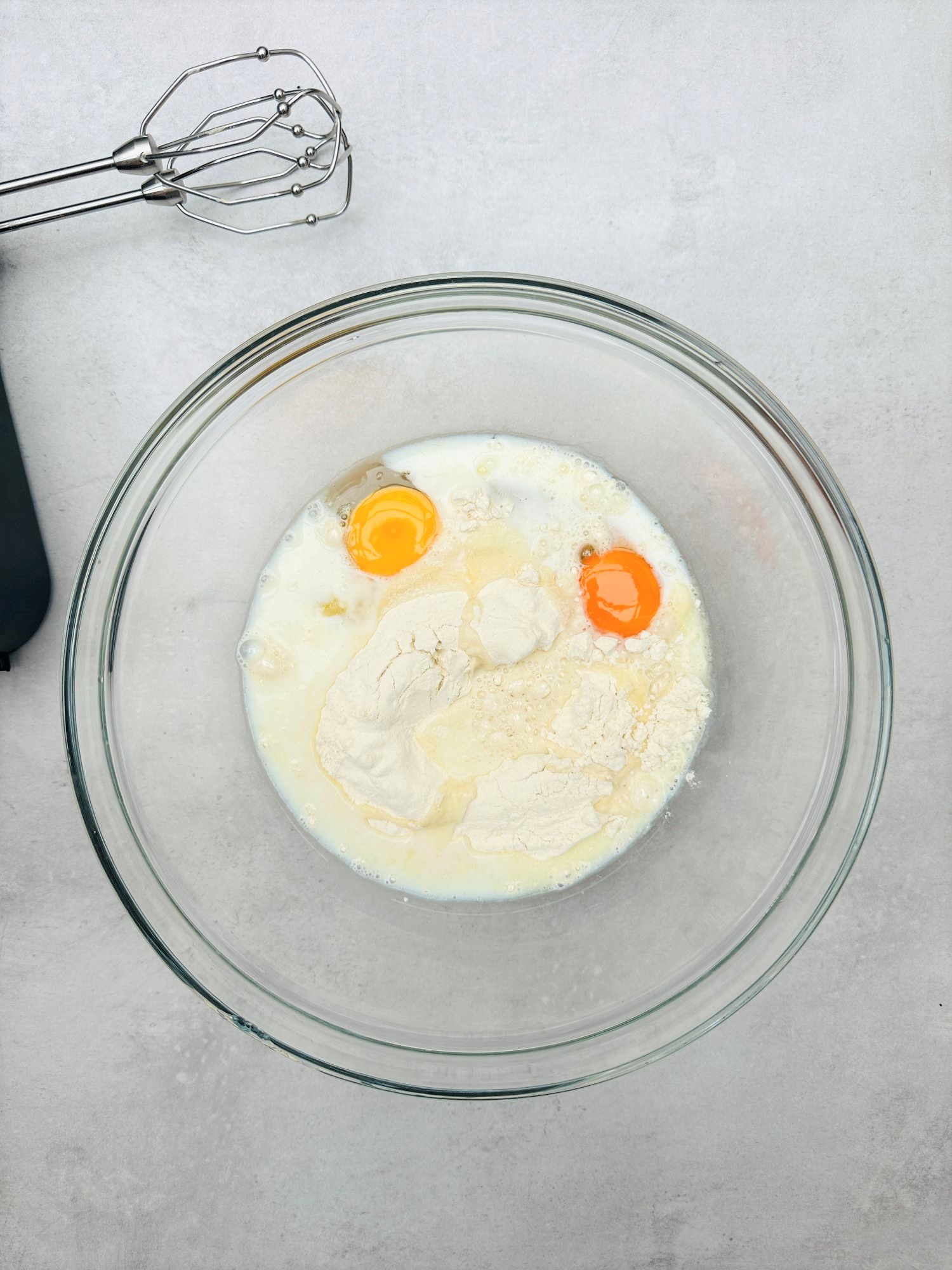 glass mixing bowl with plain flour, semi skimmed milk, two eggs with an electric whisk next to it
