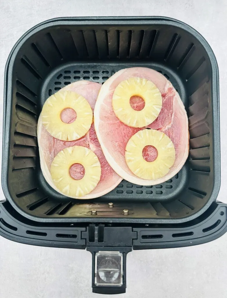 two uncooked gammon steaks in the air fryer basket with two slices of pineapple rings on each gammon steak