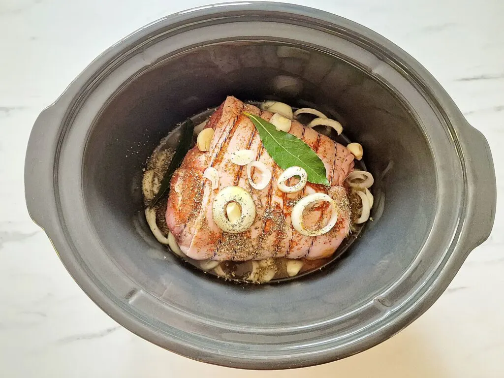 pork shoulder joint in slow cooker with hot stock, onions, bay leaves, herbs