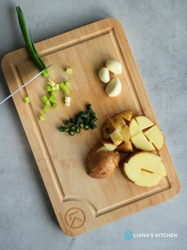 chopping ingredients to prepare them for the soup: spring onion, garlic cloves, green chillies, potato on a wooden chopping board with a Liana's Kitchen logo