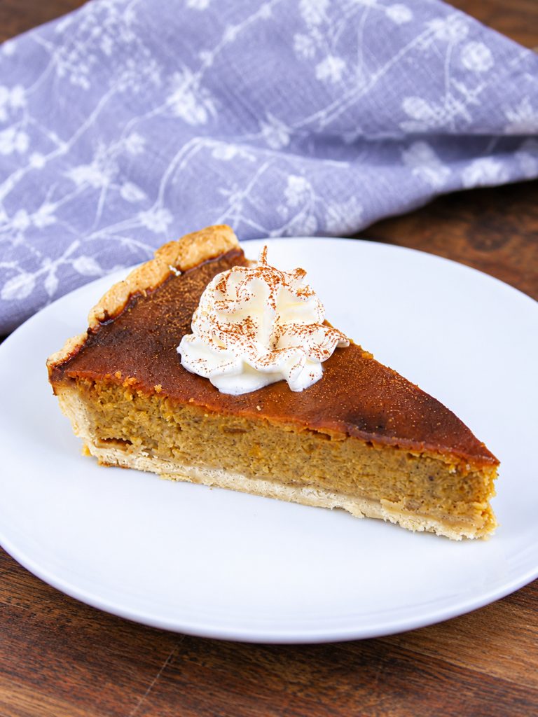 slice of pumpkin pie on a plate with cinnamon dusted across it and a squirt of fresh cream on top