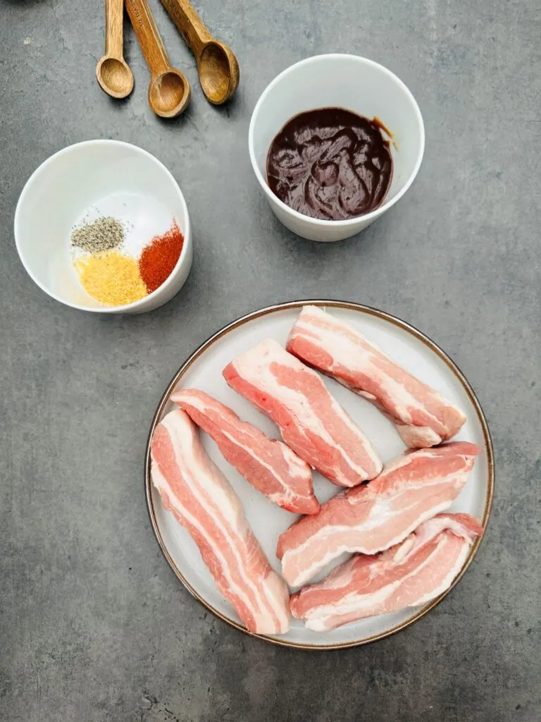 ingredients for cooking pork belly slices in an air fryer: 6 raw pork belly slices, seasoning, BBQ sauce and measuring spoons