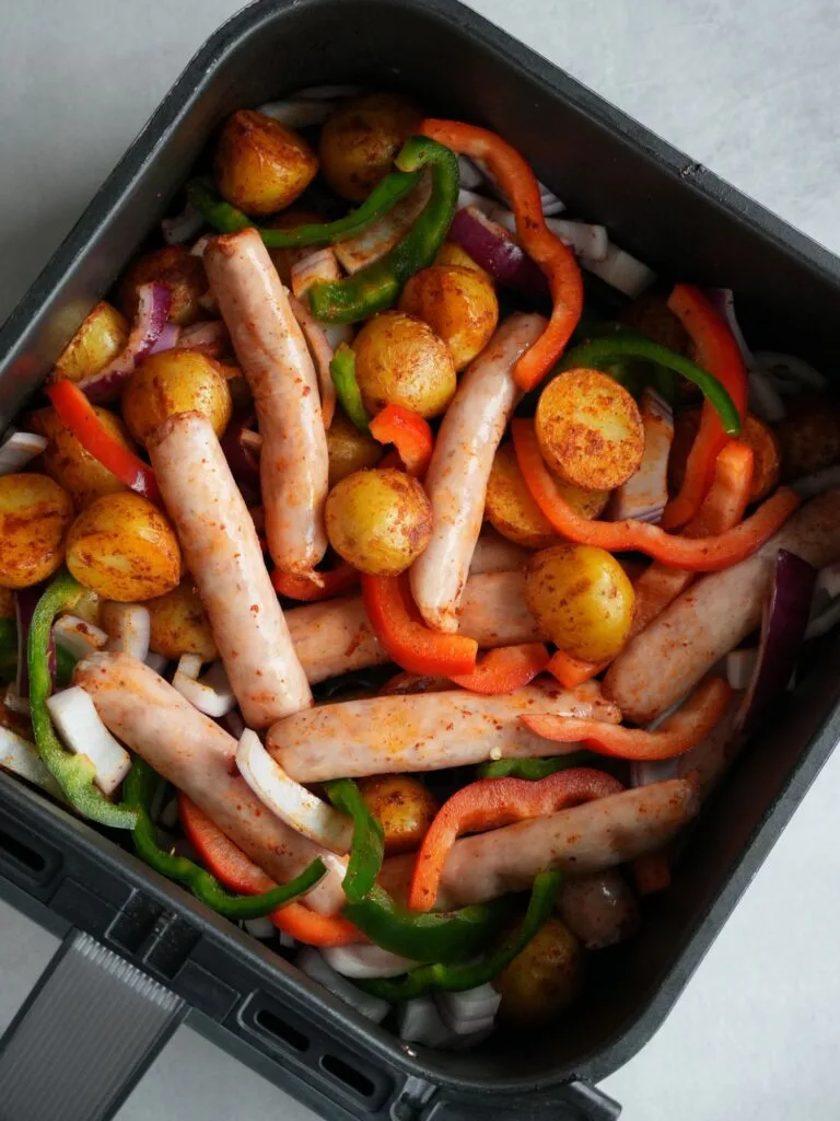 air fryer basket with raw chipolata sausages added to basket with peppers and red onions and partially cooked baby potatoes