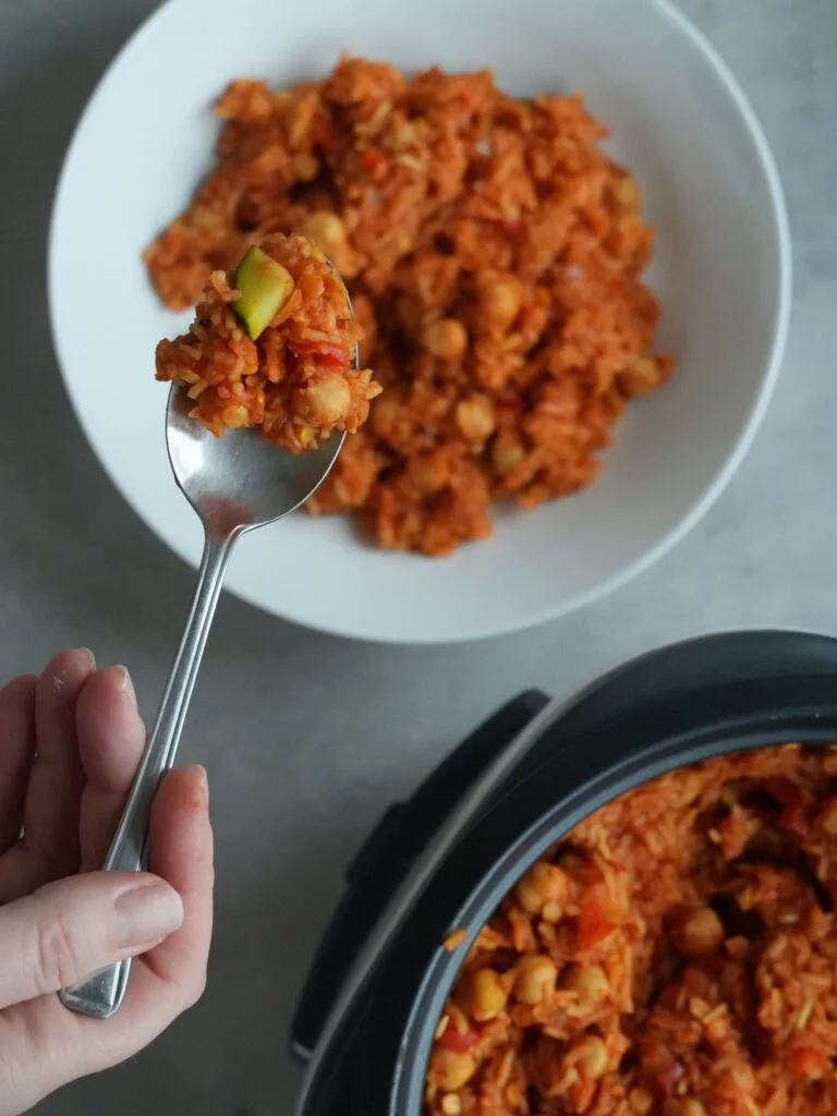 spoonful of rice on a spoon above a bowl of cooked rice, chickpeas, split red lentils and vegetables next to a rice cooker
