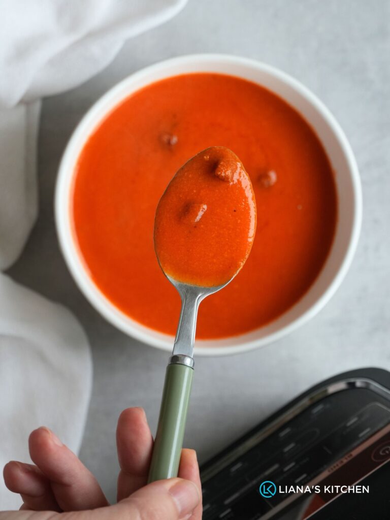 roasted pepper, chorizo and tomato soup in a white bowl with a spoon held above it, the ninja soup maker base can be seen in the corner