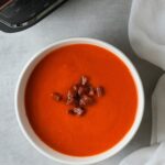 roasted red pepper, tomato and chorizo soup in a white bowl with cooked chopped chorizo sprinkled on top next to a white cloth with part of the ninja soup maker showing in the corner