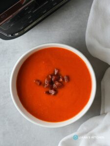 roasted red pepper, tomato and chorizo soup in a white bowl with cooked chopped chorizo sprinkled on top next to a white cloth with part of the ninja soup maker showing in the corner