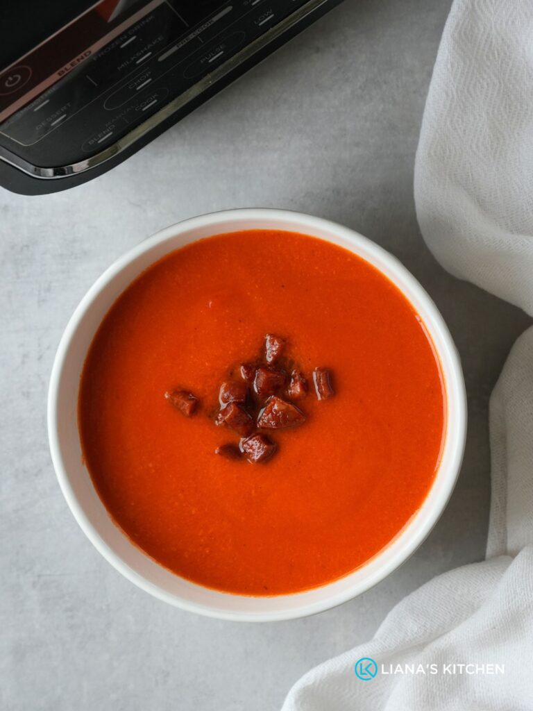 roasted red pepper, tomato and chorizo soup in a white bowl with cooked chopped chorizo sprinkled on top next to a white cloth with part of the ninja soup maker showing in the corner
