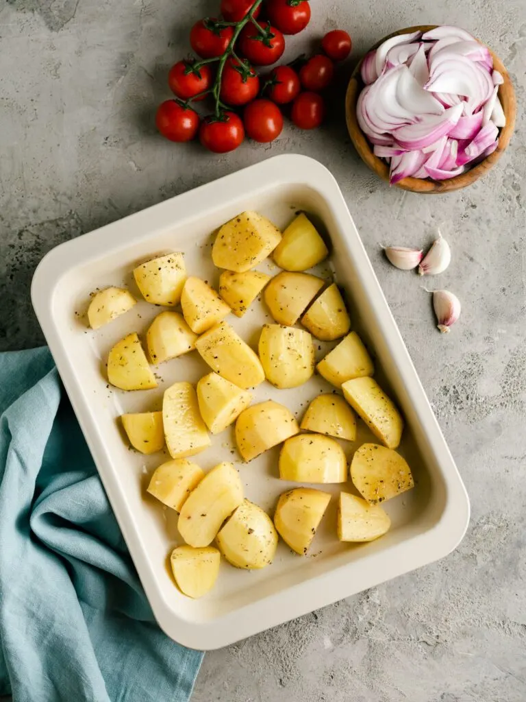 potatoes in roasting tray with cherry tomatoes, onions and garlic next to them