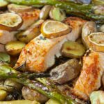 three cooked salmon fillets with lemon slices on top with asparagus spears, new potatoes and herbs in a tray bake
