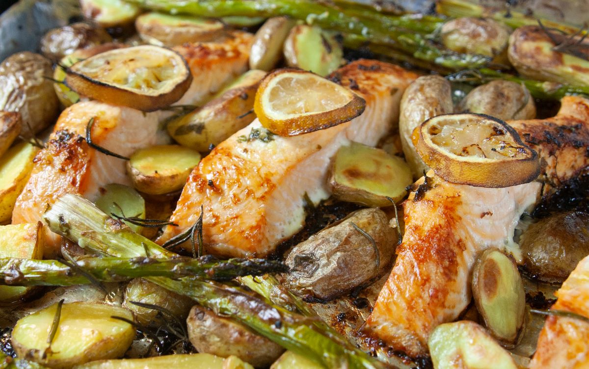 three cooked salmon fillets with lemon slices on top with asparagus spears, new potatoes and herbs in a tray bake