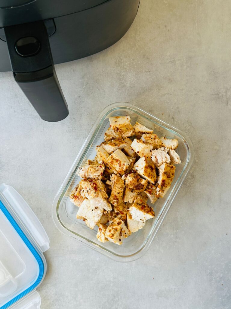 cut up cooked piri piri chicken in a glass storage container next to the lid and an air fryer