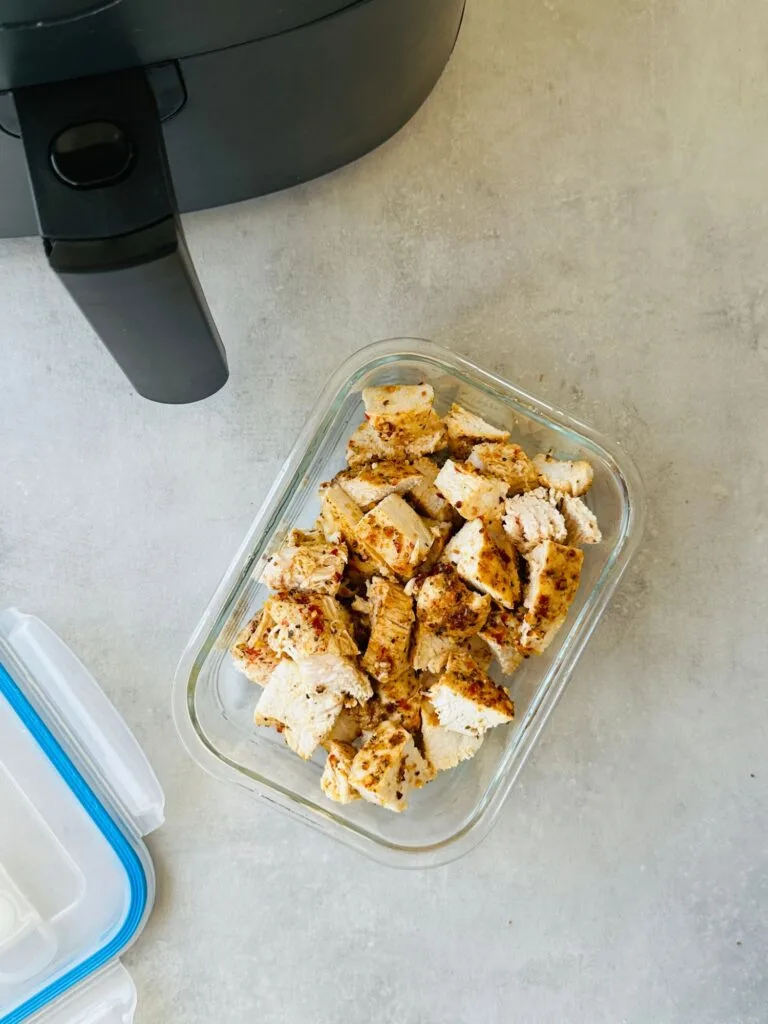 cut up cooked piri piri chicken in a glass storage container next to the lid and an air fryer