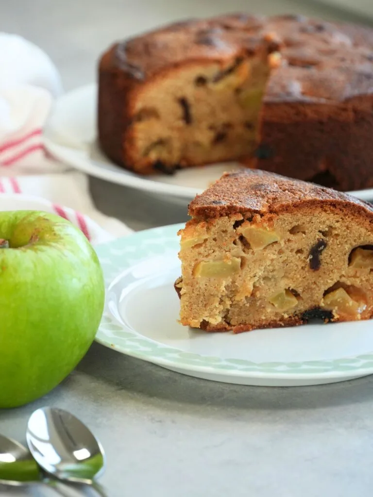 slice of apple and cinnamon cake on a plate with close up of Bramley apple next to it and the remaining cake in the background