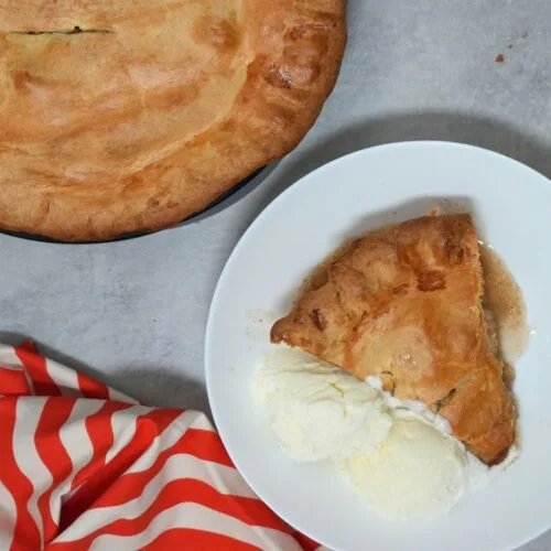 slice of apple pie in a white bowl with vanilla ice cream and a red striped cloth next to it
