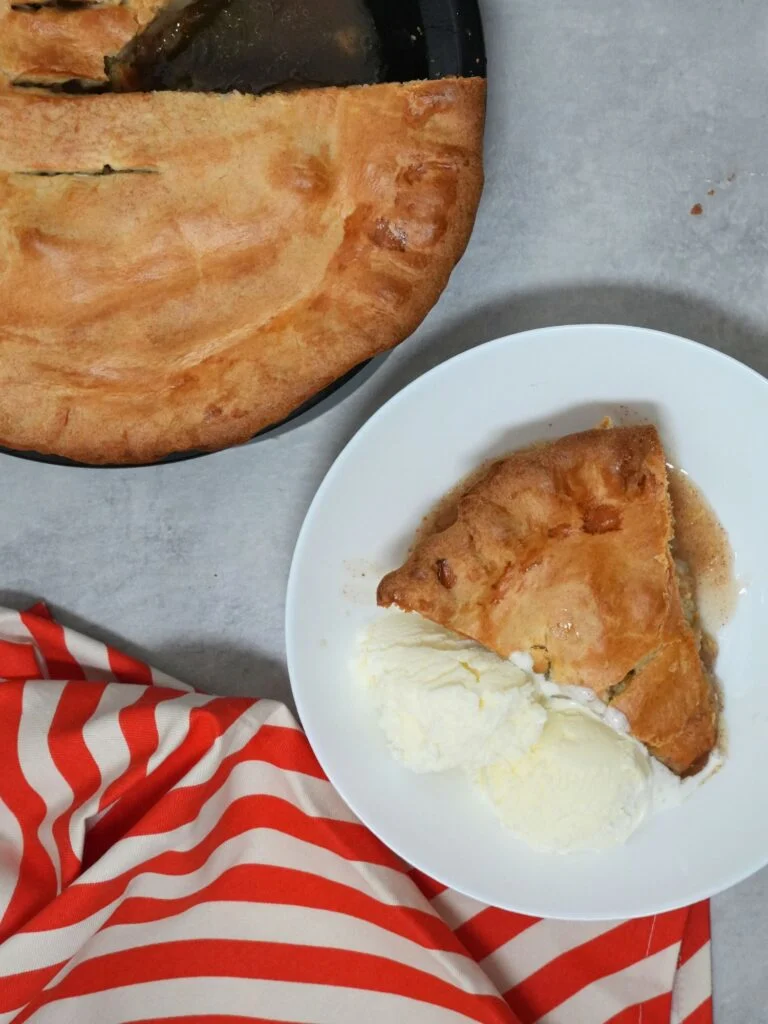 slice of apple pie in a white bowl with vanilla ice cream and a red striped cloth next to it