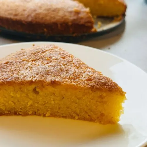 slice of lemon polenta cake on a plate in front of the remaining cake