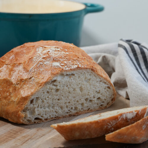 sliced no knead bread on a bread board with a knife and a dutch oven in the background
