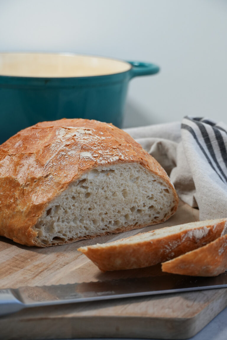 sliced no knead bread on a bread board with a knife and a dutch oven in the background