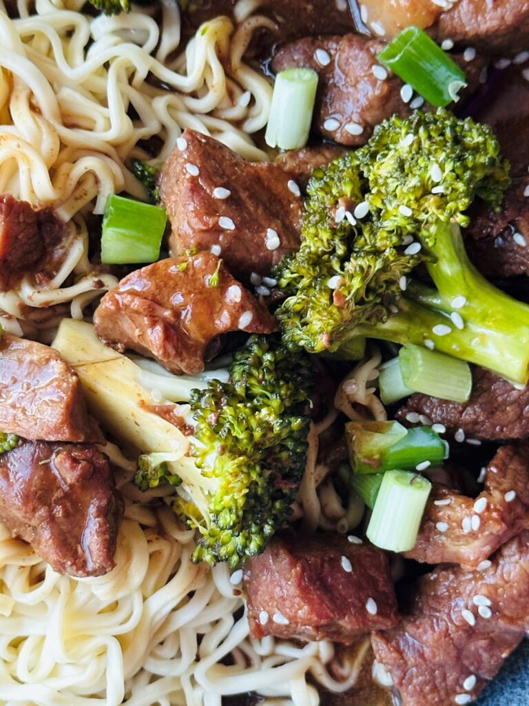 slow cooked beef and broccoli served with noodles and garnished with sesame seeds, chopped spring onions