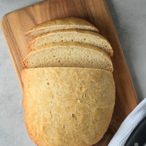 loaf of sliced bread on a wooden board with a bread knife next to it resting on a white cloth. part of a slow cooker can be seen in the top left corner.