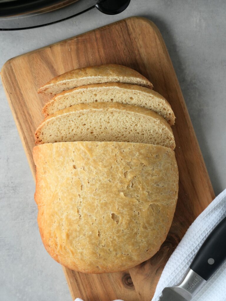 sliced bread on a wooden board with a bread knife next to it on a white cloth. part of a slow cooker can be seen in the top left corner