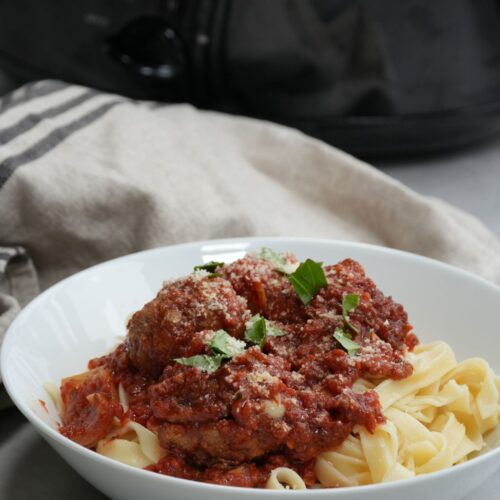 meatballs on top of tagliatelle pasta in a white bowl in front of a slow cooker