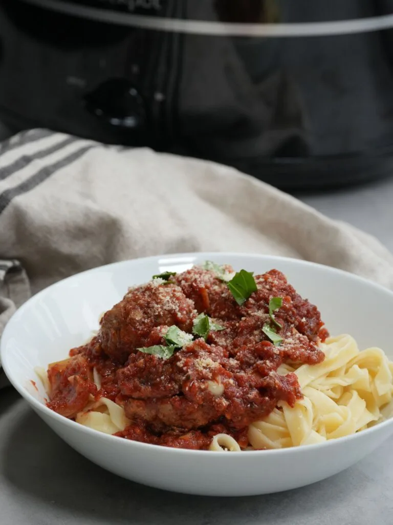 meatballs on top of tagliatelle pasta in a white bowl in front of a slow cooker