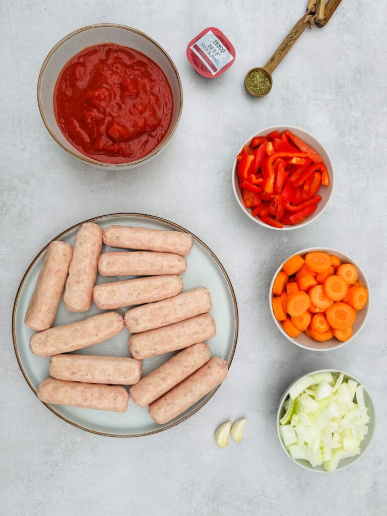 ingredients laid out for sausage casserole: 12 sausages on a plate, chopped carrots, chopped onion, chopped red pepper, 1 tsp dried mixed herbs, chopped tomatoes and beef stock pot