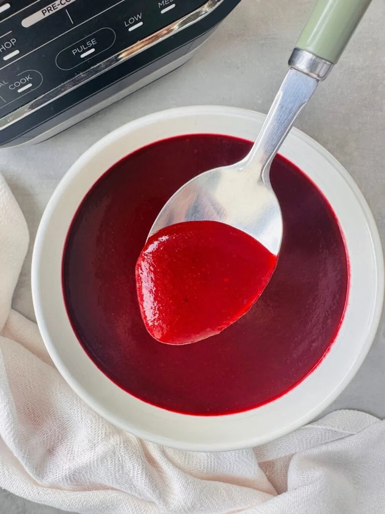 beetroot soup in a white bowl with a spoon held above it next to a white cloth and part of the ninja soup maker base in the corner