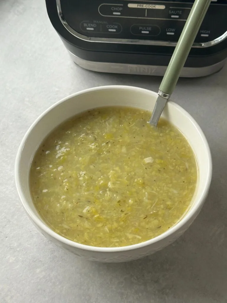 cabbage and leek soup cooked and served in a white bowl with a spoon in it in front of part of the ninja soup maker and blender