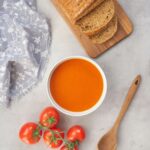 tomato soup served in a white bowl with fresh tomatoes next to it and fresh sliced bread on a chopping board