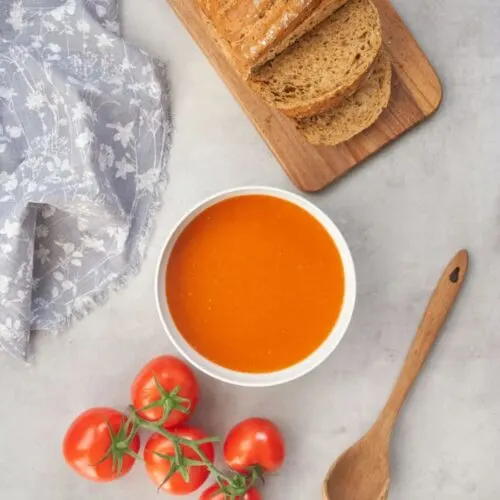 tomato soup served in a white bowl with fresh tomatoes next to it and fresh sliced bread on a chopping board