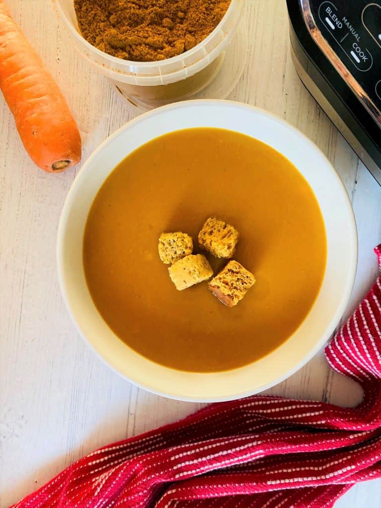 spiced carrots soup in a bowl with croutons on top next to a raw carrot and curry powder and ninja soup maker