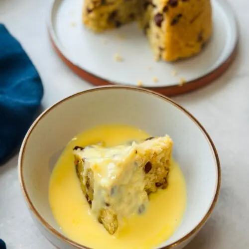 spotted dick sliced in a bowl and covered in custard with the remaining spotted dick on a plate behind