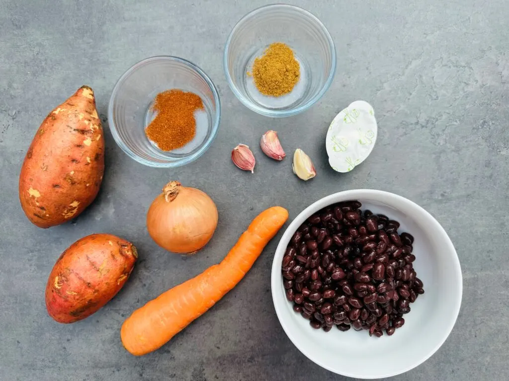 ingredients laid out for black bean and sweet potato soup: two sweet potatoes, one carrot, three cloves of garlic, one onion, vegetable stock pot, spices and black beans