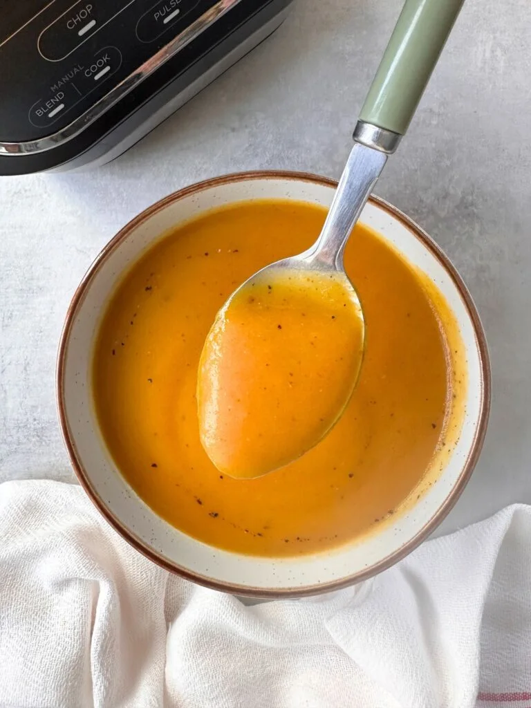 butternut squash and sweet potato soup in a bowl with a spoon held above it. part of the base of the Ninja soup maker can be seen in the corner.