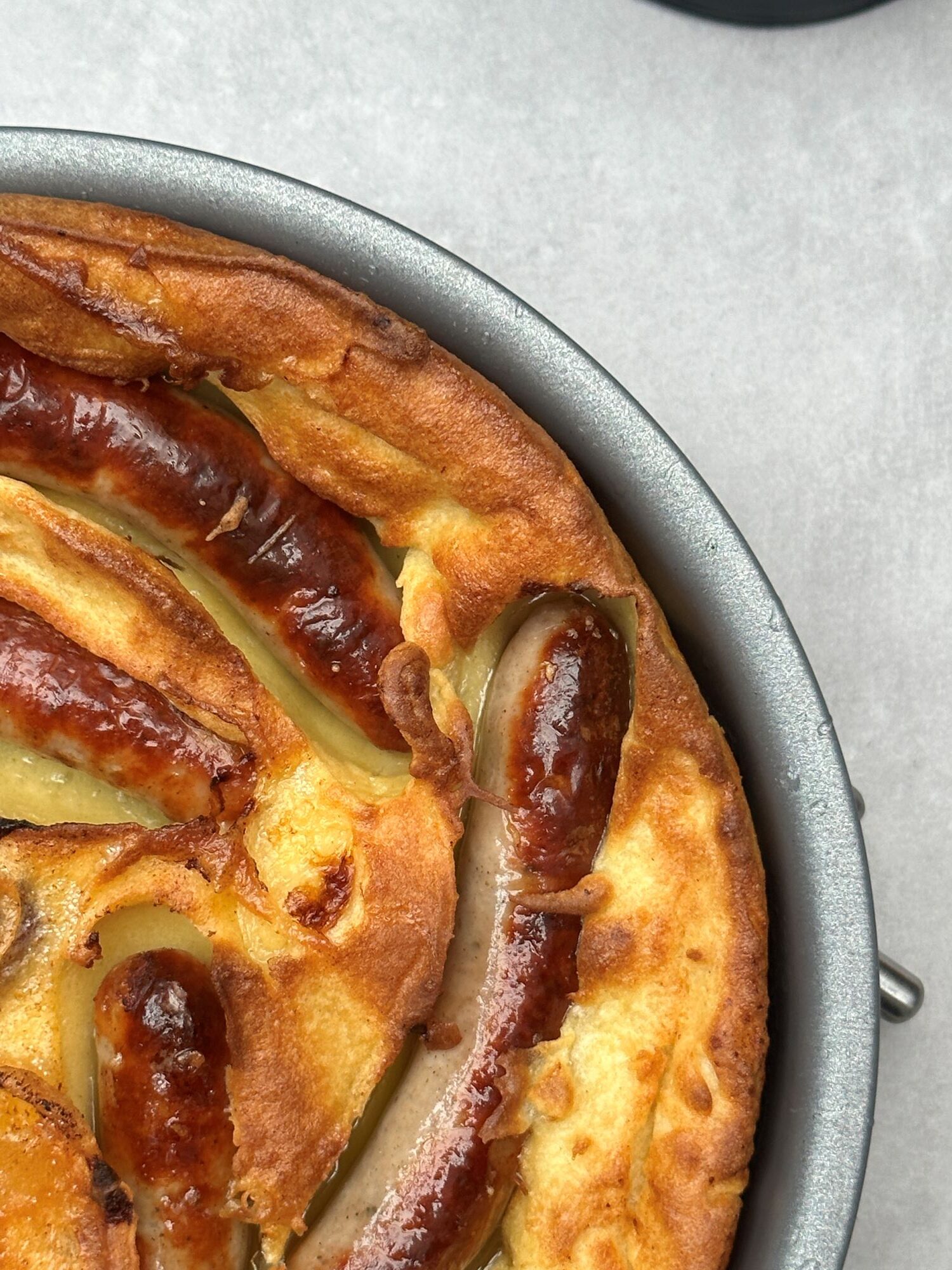 close up of toad in the hole in a baking tin. The batter has risen and the sausages are golden brown. In the corner of the picture there is part of an air fryer showing.