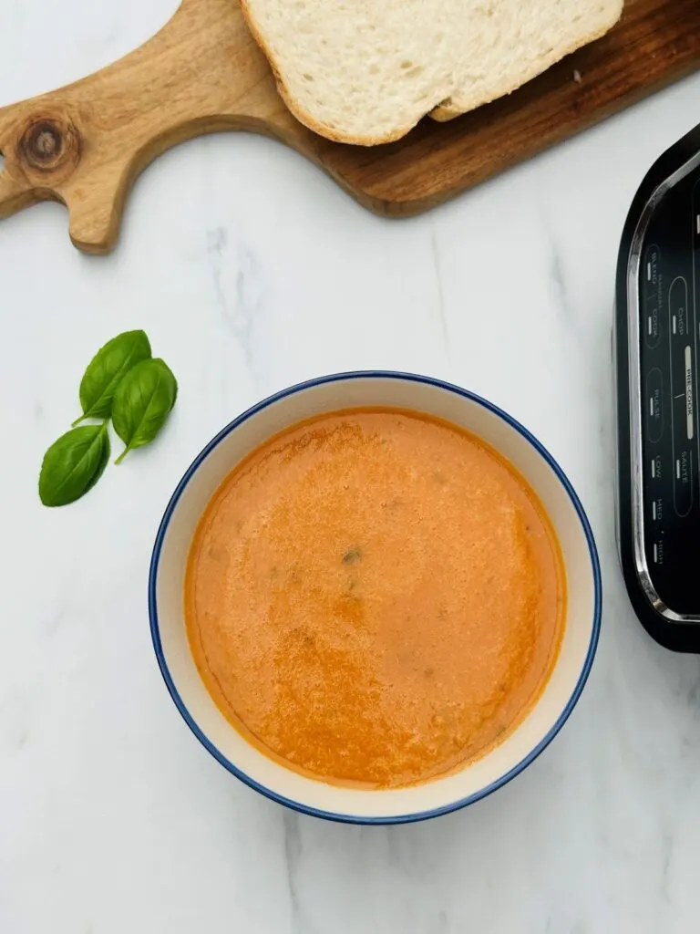 tomato with basil soup next to fresh basil leaves with Ninja soup maker next to it and bread on a chopping board