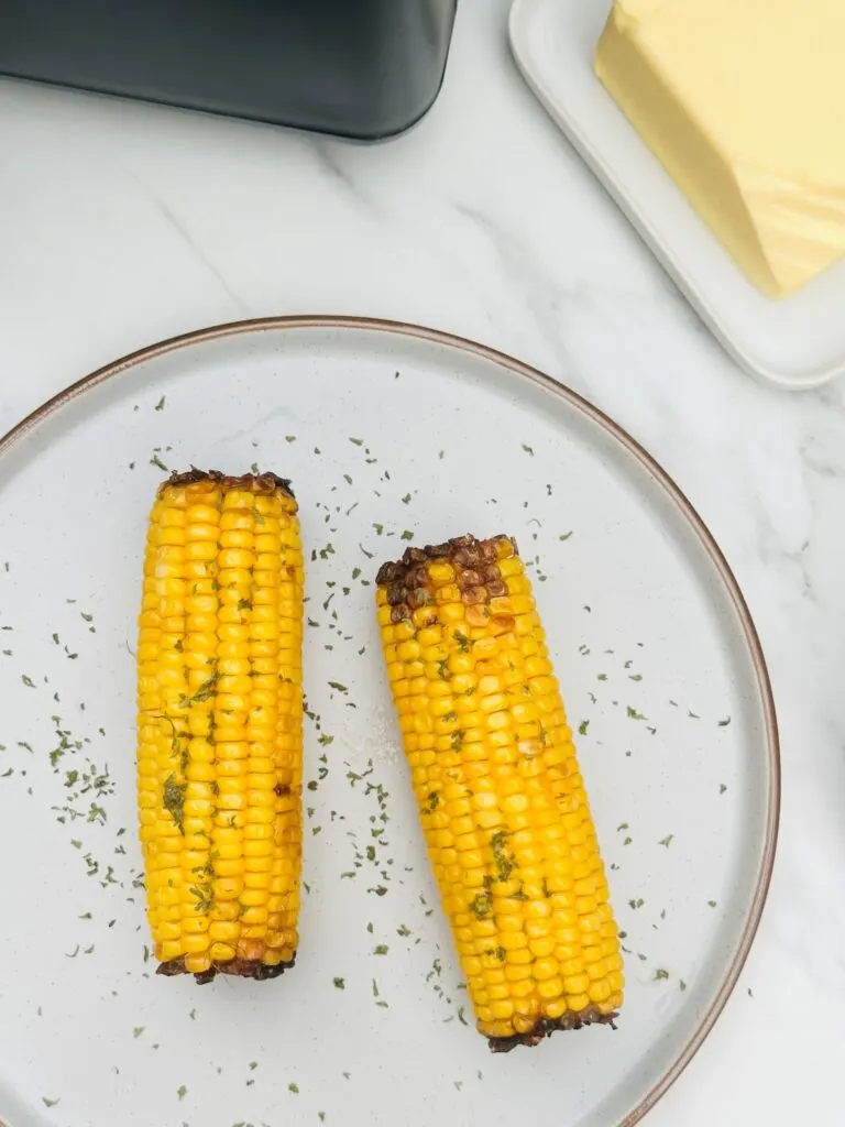 two corn on the cob next to air fryer with parsley sprinkled on top