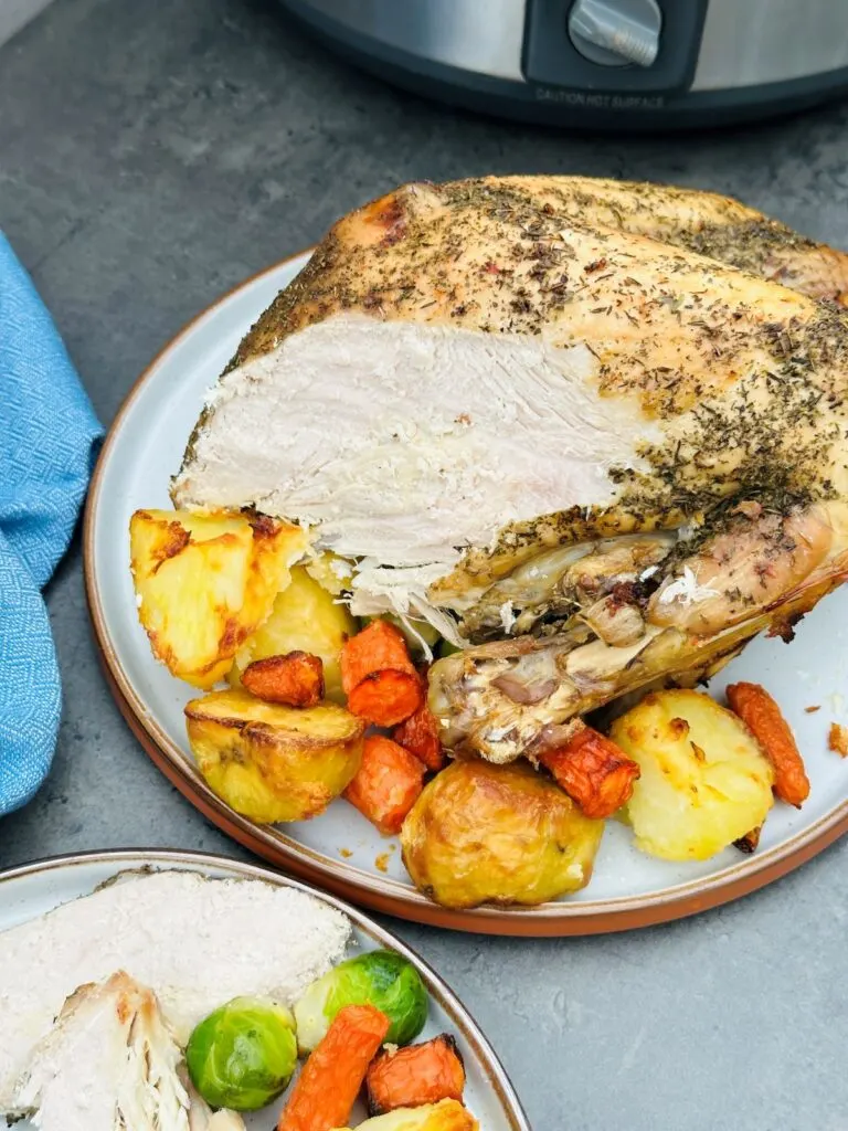 carved turkey crown on a plate with roasted potatoes, carrots and brussel sprouts next to the slow cooker it was cooked in