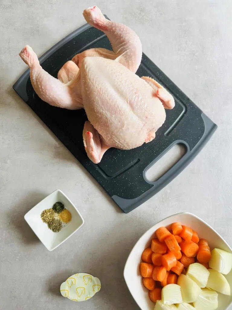 ingredients for cooking a whole chicken in a slow cooker, raw whole chicken on a chopping board, carrots, onions, herbs and spices and chicken stock pot 